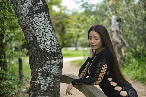 Genç bir kadın, koyu saçlı, güzel, soluk tenli, siyah bir elbise giyiyor, uzak bir bakışla, düşünceli ve endişeli, açık hava parkındaki ahşap korkuluklara yaslanmış..