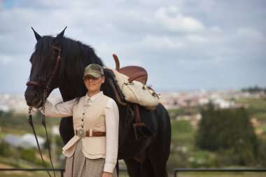 Genç bayan binici, siyah İspanyol safkan atının yanında poz veriyor, dizginleri elinde tutuyor. Konsept at biniciliği, at biniciliği, binicilik, binicilik.