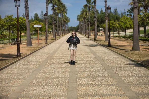 Genç, güzel, sarışın, deri ceketli, yılan desenli mini etekli, çizmeli ve güneş gözlüklü, elleri cebinde, yalnız ve bağımsız bir şekilde bir caddenin ortasında poz veriyor..