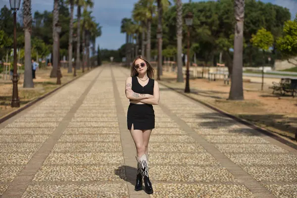 Genç, güzel, sarışın, siyah bluzlu, etekli, çizmeli ve güneş gözlüklü, kolları çapraz, güçlü ve bağımsız, bir caddenin ortasında. Kadınlık kavramı, güçlenme..