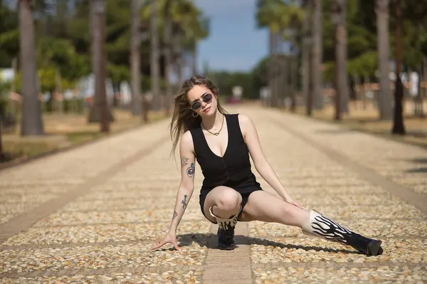 Genç, güzel bir kadın, sarışın, siyah bir bluz ve etek giyiyor, çizmeler ve güneş gözlüğü takıyor, bir bacağı esnemiş, çekici ve seksi bir şekilde bir caddenin ortasında çömelmiş poz veriyor. Güzellik kavramı.