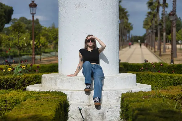 Genç bir kadın, güzel, sarışın, siyah bir tişört, kot pantolon ve güneş gözlüğü giyiyor, beyaz basamaklarda oturuyor, güneşli bir günde elini güneşten koruyor. Kavramsal sıcaklık, güneş, yaz.