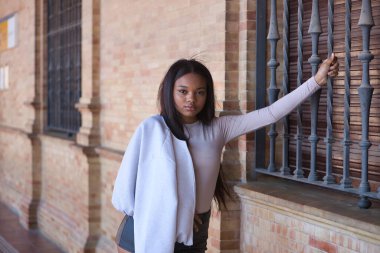 Genç, güzel, deri ceketli ve şortlu Afrikalı bir kadın, kameraya ciddi bir hareketle bakıyor, Seville 'de İspanya meydanında bir pencerenin ızgarasına tutunuyor..