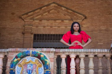 Genç, güzel, kırmızı elbiseli Afrikalı bir kadın Seville 'de İspanya meydanında elleri belinde kameraya bakıp mutlu mesut gülümsüyor. Moda konsepti, güzellik, moda.