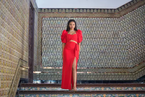 Genç, güzel, Afrikalı bir kadın, kırmızı bir elbise giyiyor, Sevilla 'daki İspanyol meydanının merdivenlerinde mankenlik yapıyor. Moda konsepti, güzellik, moda.