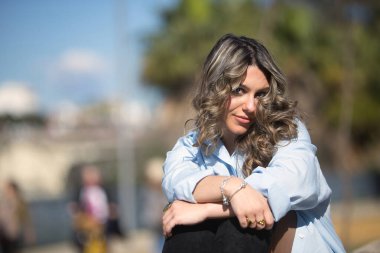 Genç, güzel, sarışın kadın, gömlek, deri etek ve botlar giyiyor, kameraya bakıyor, rahat ve sakin, taş bir duvarda oturuyor..