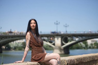 Genç bir kadın, esmer, uzun boylu, zayıf ve çok dövmeli, kahverengi elbiseli, arkasında Triana köprüsü olan nehir kenarında sonsuzluğa bakıyor. Gençlik kavramı, bin yıllık.