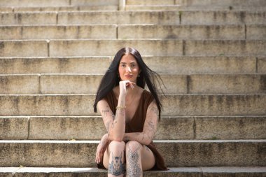 Genç bir kadın, koyu saçlı, uzun boylu, zayıf ve çok dövmeli, kahverengi bir elbise giyiyor, eli çenesinde, kameraya bakıyor, tatlı, masum ve yalnız, merdivenlerde oturuyor..