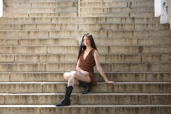 Genç bir kadın, esmer, uzun boylu, zayıf ve çok dövmeli, kahverengi elbiseli ve çizmeli, tatlı, masum ve yalnız, buharlaşmış, merdivenlerde oturuyor. Gençlik kavramı, güzellik, asilik, milenyum.