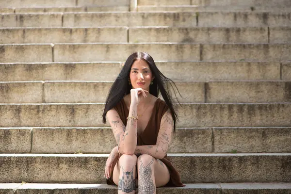 Genç bir kadın, koyu saçlı, uzun boylu, zayıf ve çok dövmeli, kahverengi bir elbise giyiyor, eli çenesinde, kameraya bakıyor, tatlı, masum ve yalnız, merdivenlerde oturuyor..
