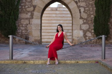 Genç, güzel, esmer, güzel kırmızı parti elbisesi içinde gülümseyen, mutlu ve komik poz veren bir kadın sokakta büyük bir zincirde oturuyor. Güzellik, moda, moda, gençlik kavramı.