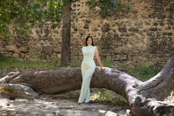 Beyaz elbiseli, genç, güzel, esmer kadın devrilmiş bir ağacın gövdesine yaslanmış kameraya bakıyor. Güzellik, moda, moda, gençlik kavramı.