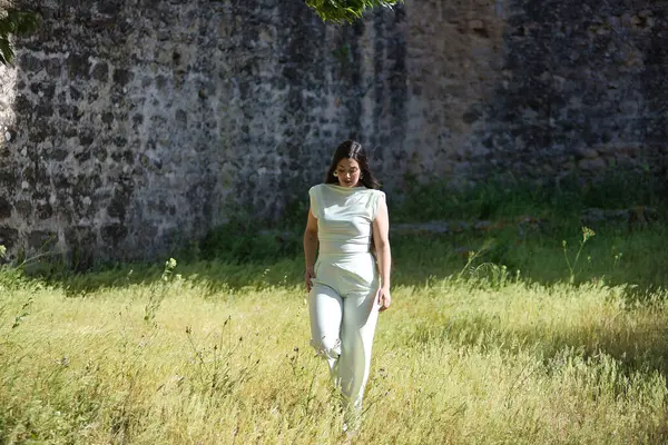 Beyaz elbiseli, genç, güzel, esmer bir kadın, rahat ve sakin, güneşli bir bahar gününde uzun çimlerde yürüyor. Güzellik, moda, moda, gençlik kavramı.
