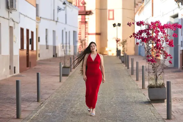 Genç, güzel, esmer bir kadın, güzel kırmızı bir parti elbisesi giyiyor, Endülüs köyünün güzel ve yalnız bir caddesinde yürüyor. Güzellik, moda, moda, gençlik kavramı.