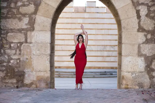 Genç, güzel, esmer, güzel kırmızı parti elbisesi ve topuklu ayakkabıları olan, kameraya bakan, kolları yukarıda taş kemerin ortasında duran bir kadın. Güzellik, moda, moda, gençlik kavramı.