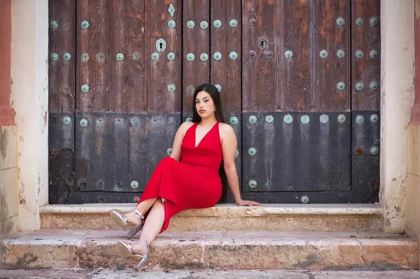 Genç, güzel, esmer bir kadın, güzel kırmızı bir parti elbisesi ve topuklu ayakkabıları içinde, rahat ve sakin, bazı basamaklarda oturuyor ve eski bir ahşap kapıya yaslanıyor. Güzellik, moda, moda, gençlik kavramı.