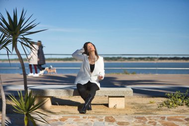 Genç bir kadın, güzel, kahverengi saçlı, beyaz ceketli, gözleri kapalı, eli saçında, bankta oturuyor, yalnız ve sessiz, şafakta güneş ışınlarını alıyor..