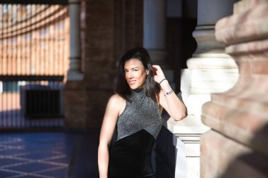 Genç, güzel, esmer bir kadının portresi, süslü siyah bir elbise giyiyor, kameraya bakıyor, rahat ve sakin, mermer bir sütuna yaslanıyor, güneş ışınlarını alıyor. Güzellik kavramı, kadınlık.