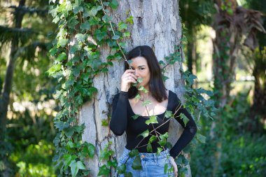 Genç, güzel, esmer bir kadın, siyah tişört ve kot pantolon, bir ağacın gövdesine yaslanmış, sarmaşıkla komik oynuyor, doğayla bağlantılı. Güzellik kavramı, kadınlık, doğa, bitkiler, ağaçlar.