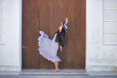 Genç bir kadın, güzel, esmer, flamenko dansçısı, ceketli ve şapkalı güzel bir elbise giyiyor, arka planda ahşap kapılı flamenko dansı yapıyor. Flamenko konsepti, tipik, İspanyolca, Sevilla.