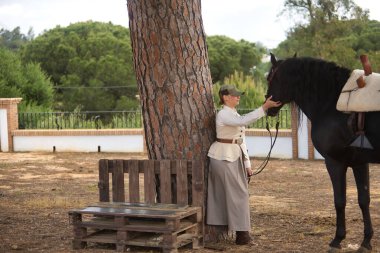 Genç kadın binici bir ağaca yaslanıyor, siyah İspanyol safkan atını okşuyor, sevgisini ve şefkatini gösteriyor. Konsept at biniciliği, at biniciliği, binicilik, binicilik.