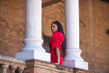 Genç, güzel, kırmızı elbiseli Afrikalı bir kadın. İspanya 'nın Sevilla meydanındaki iki mermer sütunun arasında sonsuzluğa bakıyor. Moda konsepti, güzellik, moda.