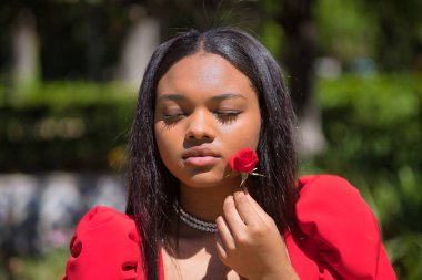 Genç, güzel, kırmızı elbiseli Afrikalı bir kadının portresi, kırmızı bir gül ile yüzünü okşuyor, tatlı, hassas ve kırılgan. Güzelliği kavra, çiçekleri, baharı.