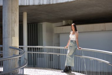 Genç bir kadın, esmer, uzun boylu, zayıf ve çok dövmeli, beyaz bluzlu, kot etekli ve botlu, kameraya bakıyor, saçlarına güneş ışınları tutuyor, korkuluklara yaslanıyor..