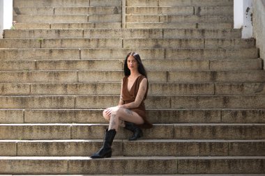 Genç bir kadın, esmer, uzun boylu, zayıf ve çok dövmeli, kahverengi elbiseli ve çizmeli, tatlı, masum ve yalnız, merdivenlerde oturuyor. Gençlik kavramı, güzellik, asilik, milenyum.