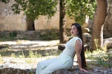 Beyaz elbiseli, genç, güzel, esmer bir kadın, rahat ve sakin, büyük bir kayanın üzerinde oturan kameraya bakıyor. Güzellik, moda, moda, gençlik kavramı.