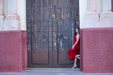 Genç, güzel, esmer, güzel bir kadın güzel kırmızı bir parti elbisesi ve topuklu ayakkabıları içinde, eski bir kapının çerçevesine yaslanmış. Güzellik, moda, moda, gençlik kavramı.