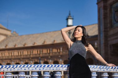 Genç, güzel, esmer, süslü siyah elbiseli bir kadın kameraya bakıyor, Seville 'de İspanya meydanındaki bir köprüde fayanslı bir korkuluğa yaslanıyor. Güzelliği, kadınlığı, turizmi kavrayın.