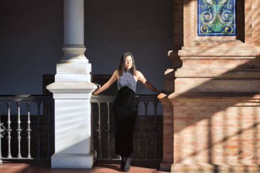 Genç, güzel, esmer bir kadın, siyah süslü bir elbise giymiş, gözleri kapalı, korkuluklara yaslanmış, güneşin ışınlarını alan. Güzelliği, modayı, modayı, kadınlığı kavra.