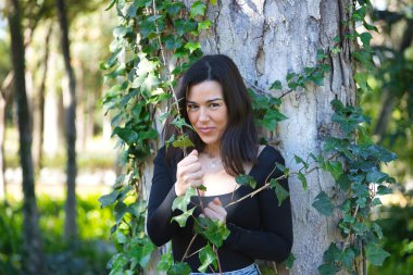Siyah tişört ve kot pantolon giymiş, bir ağacın gövdesine yaslanmış ve doğayla bağlantılı, sarmaşığa dolanmış genç, güzel, koyu renk saçlı bir kadın. Güzellik kavramı, kadınlık, doğa, bitkiler, ağaçlar.