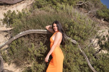 Genç, güzel, turuncu elbiseli, kumsalda ağaç köklerinin yanında saçlarına dokunan esmer bir kadın. Güzellik, doğa, görünüş, moda kavramı.