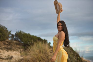 Sarı elbiseli, genç, güzel, koyu renk saçlı bir kadın, bir kolu havada, elinde şapkasıyla, gülümseyen ve eğlenen kameraya bakıyor. Güzelliği, modayı, eğlenceyi, mutluluğu, hayatı kavra.