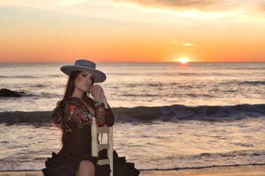 Kadın, genç, güzel, esmer, flamenko dansçısı, kordova şapkalı ve çiçekli siyah flamenko elbiseli, plajda arka planda deniz olan bir sandalyede oturuyor..