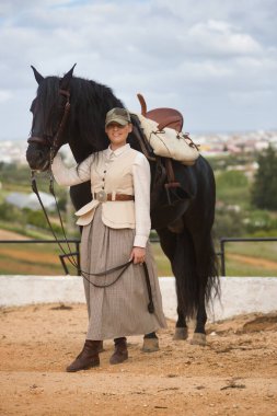 Genç bayan binici, siyah İspanyol safkan atının yanında poz veriyor, dizginleri elinde tutuyor. Konsept at biniciliği, atlı kadın, at biniciliği, binicilik. Dikey konum.