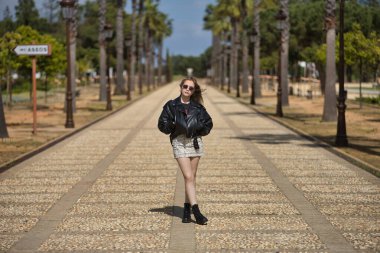 Genç, güzel, sarışın, deri ceketli, yılan desenli mini etekli, çizmeli ve güneş gözlüklü, elleri cebinde, yalnız ve bağımsız bir şekilde bir caddenin ortasında poz veriyor..