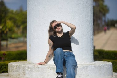 Genç bir kadın, güzel, sarışın, siyah bir tişört, kot pantolon ve güneş gözlüğü giyiyor, beyaz basamaklarda oturuyor, güneşli bir günde elini güneşten koruyor. Kavramsal sıcaklık, güneş, yaz.