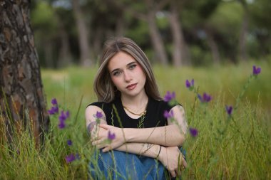 Genç bir kadın, güzel, sarışın, yeşil gözlü, siyah tişört ve kot pantolon giyen, tatlı ve narin, çimenlerde oturan ve mor çiçeklerle çevrili. Masumiyet kavramı, çiçekler, bahar, ot.