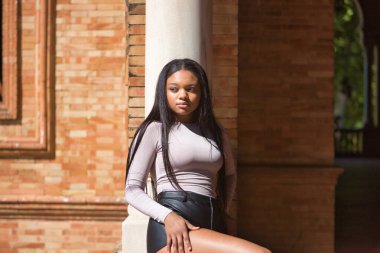 Genç, güzel, Afrikalı bir kadın, süveter ve deri şort giyiyor, sonsuzluğa bakıyor, Sevilla 'da İspanya meydanında bir korkulukta oturuyor. Moda konsepti, güzellik, moda.