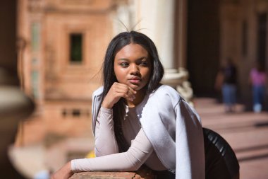 Genç, güzel, Afrikalı bir kadının portresi, deri ceket ve şort giymiş, eli çenesinde ve bakışları kaymış Seville 'de İspanya meydanında bir korkuluğa yaslanmış..