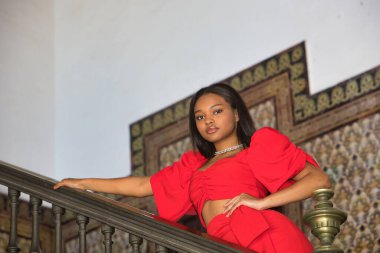 Genç, güzel, Afrikalı bir kadın, kırmızı bir elbise giyiyor, model gibi poz veriyor, Seville 'de İspanya meydanının korkuluklarına yaslanmış kameraya bakıyor. Moda konsepti, güzellik, moda.