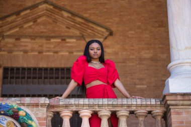 Genç, güzel, kırmızı elbiseli Afrikalı bir kadın, Seville 'de İspanya meydanındaki korkuluklara yaslanan kameraya ciddi bir jest yapıyor. Moda konsepti, güzellik, moda.