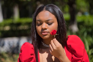 Genç, güzel, kırmızı elbiseli Afrikalı bir kadının portresi, kırmızı bir gül ile yüzünü okşuyor, tatlı, hassas ve kırılgan. Güzelliği kavra, çiçekleri, baharı.