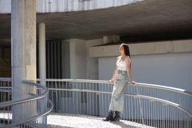 Genç bir kadın, esmer, uzun boylu, zayıf ve çok dövmeli, beyaz bluzu, kot eteği ve botları var, gözleri kapalı, saçlarına güneş ışınları alıyor, korkuluklara yaslanıyor. Gençlik kavramı, güzellik.