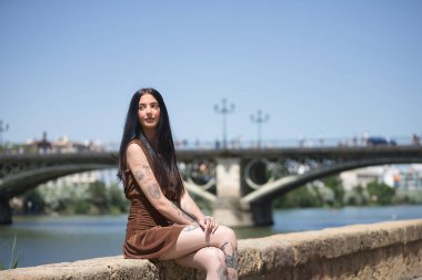 Genç bir kadın, esmer, uzun boylu, zayıf ve çok dövmeli, kahverengi elbiseli, arkasında Triana köprüsü olan nehir kenarında sonsuzluğa bakıyor. Gençlik kavramı, bin yıllık.