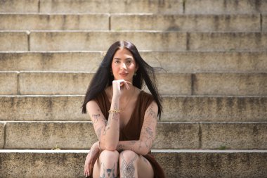 Genç bir kadın, koyu saçlı, uzun boylu, zayıf ve çok dövmeli, kahverengi bir elbise giyiyor, eli çenesinde, kameraya bakıyor, tatlı, masum ve yalnız, merdivenlerde oturuyor..