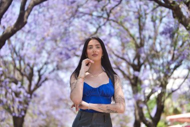 Genç bir kadın, koyu saçlı, uzun boylu, zayıf ve ağır dövmeli, mavi bluzlu ve deri şortlu, eli çenesinde ve mor bougainvillea ağaçlarıyla çevrili kameraya bakıyor..